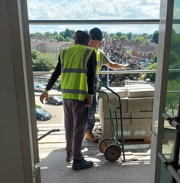 Two members of the team in high vis jackets move a trolley loaded with concrete heavy duty bricks outside on an upper floor of a commercial building.